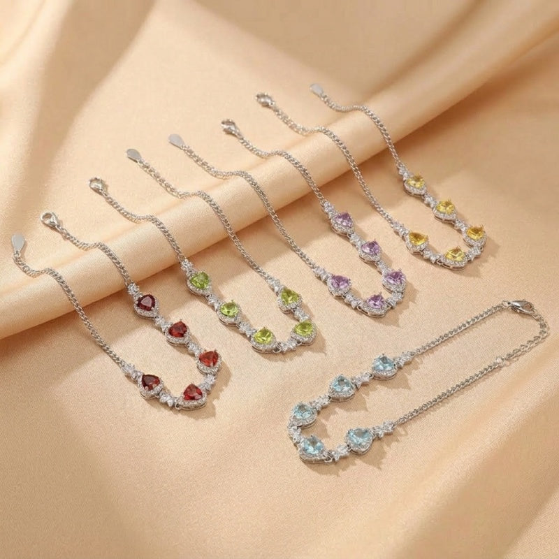 Collection de bracelets en argent avec des pierres précieuses en forme de cœur : grenat rouge, péridot vert, améthyste violette, citrine jaune et topaze bleue, sur fond satiné doré.