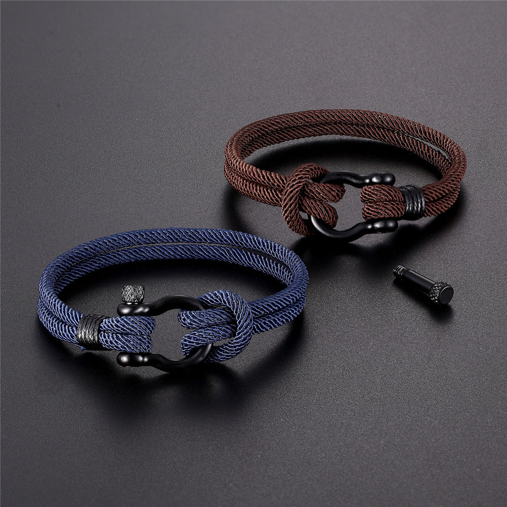 Deux bracelets en corde tressée, un bleu marine et un marron, avec fermoir mousqueton noir mat. Un petit cylindre noir est posé à côté.