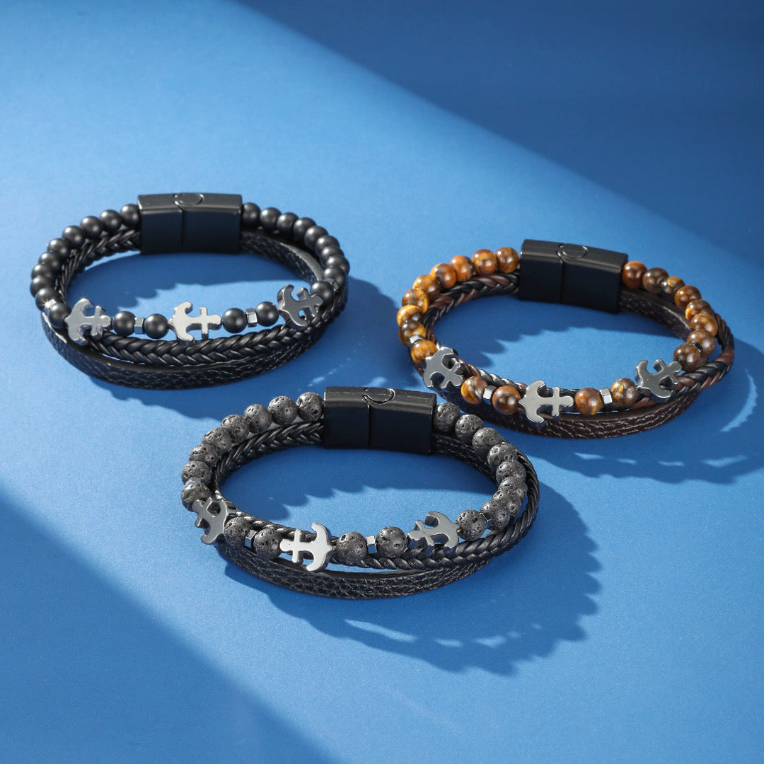 Trois bracelets pour hommes en cuir et perles avec breloques ancre et fermoir magnétique, sur fond bleu.