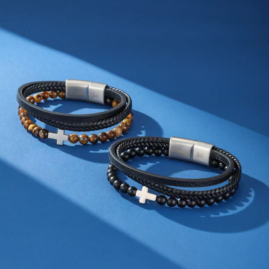 Deux bracelets pour hommes, l'un avec des perles œil de tigre et cuir noir, l'autre avec des perles noires et cuir tressé, tous deux ornés d'une croix argentée et d'un fermoir magnétique.