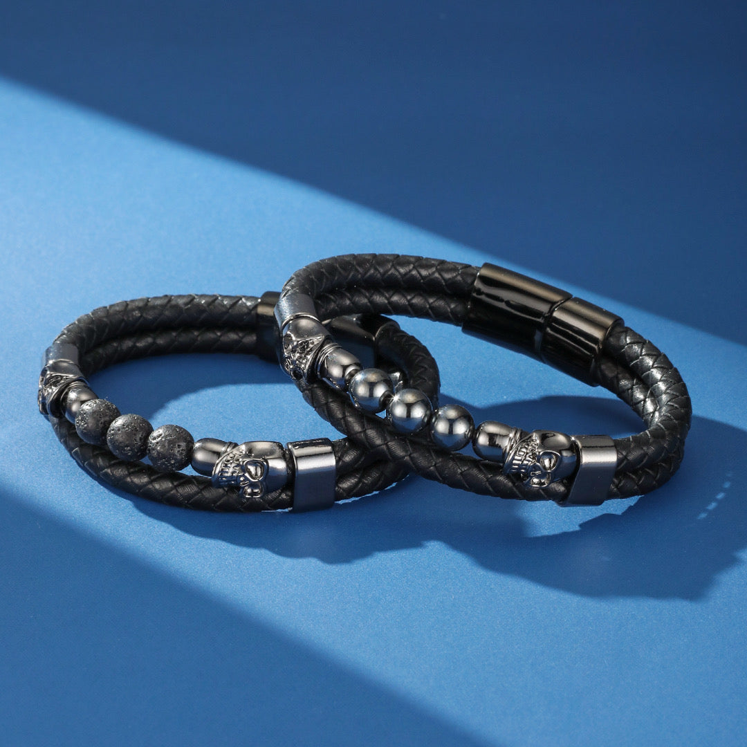 Deux bracelets masculins en cuir tressé noir, ornés de perles de pierre de lave ou d'hématite et d'éléments en forme de crâne, sur fond bleu.