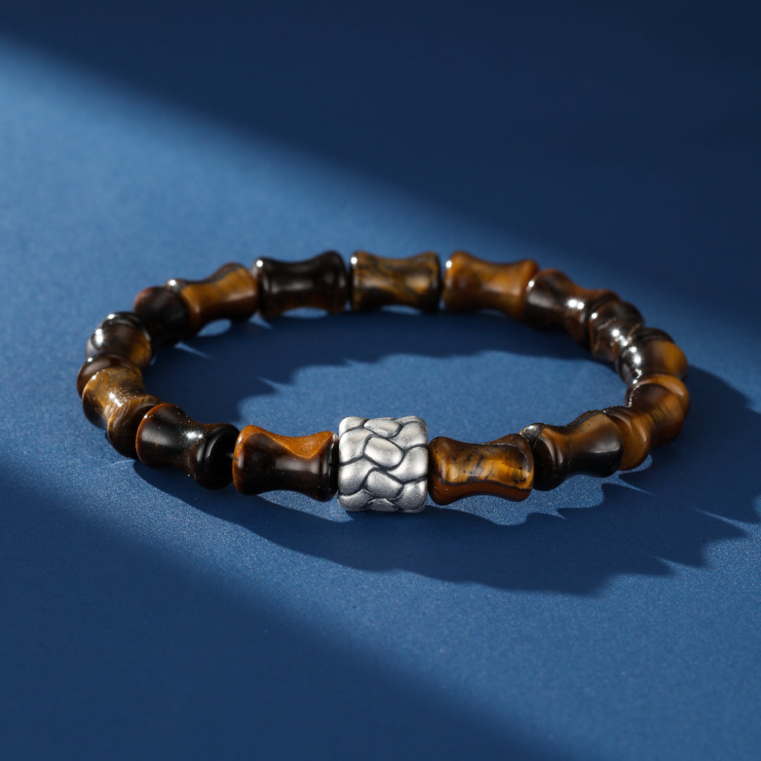 Bracelet homme en œil de tigre avec des perles en forme de bambou et un fermoir central en argent texturé sur fond bleu.