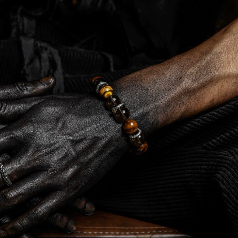 Bracelet pour homme avec des perles d'œil de tigre et des perles sombres, monté sur un poignet à la peau peinte en noir.