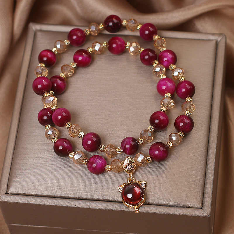 Bracelet double rang avec perles rondes fuchsia et cristaux dorés, orné d'un pendentif tête de chat avec une pierre rouge.