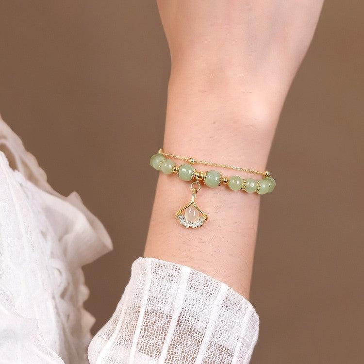 Bracelet double chaîne en jade vert, perles dorées et breloque feuille de ginkgo ornée de cristaux, porté au poignet d'une femme vêtue de blanc.