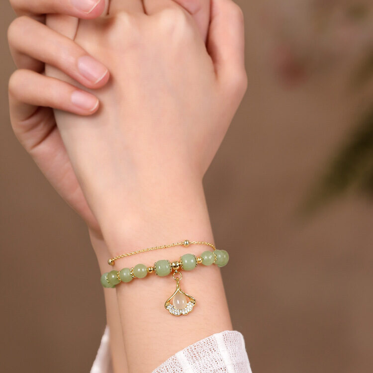 Bracelet double chaîne avec perles de jade vert, breloque feuille de ginkgo plaqué or sertie de cristaux et une pierre nacrée au centre, porté au poignet d'une femme.