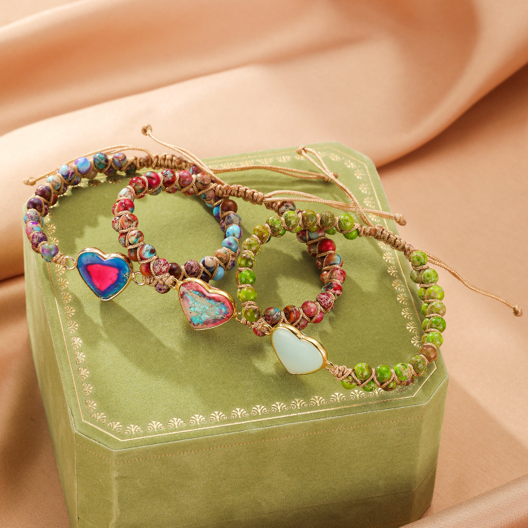 Trois bracelets en jaspe avec des pendentifs en forme de cœur de différentes couleurs, sur un fond vert satiné.