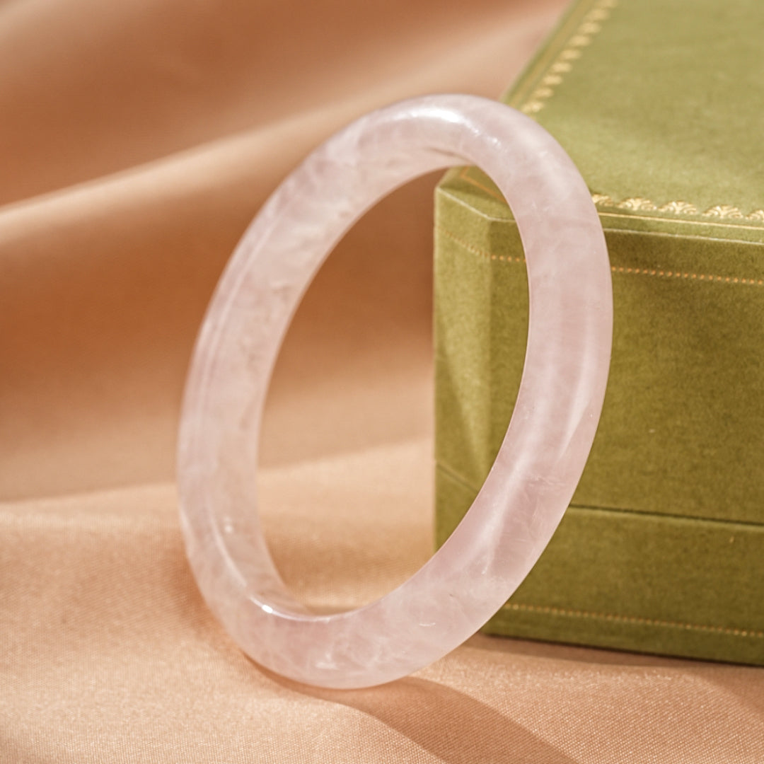 Bracelet jonc en quartz rose translucide, posé à côté d'une boîte verte sur un tissu satiné couleur pêche.