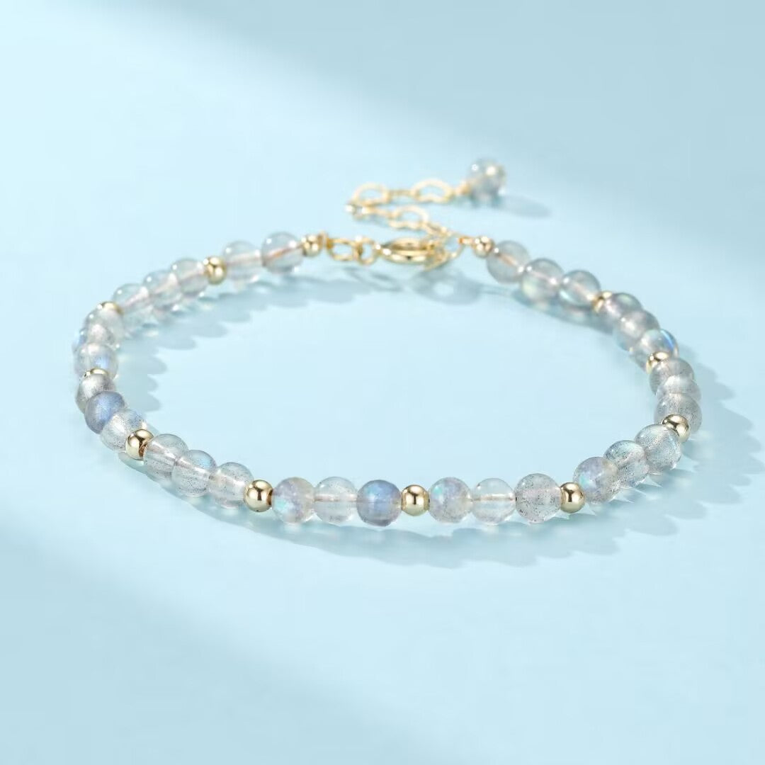 Bracelet fin en labradorite naturelle avec des perles dorées, sur fond bleu pastel.