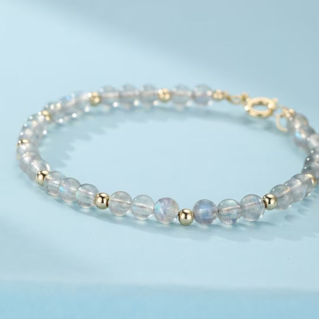 Bracelet fin en pierres naturelles de labradorite bleutées et grises, alternées avec de petites perles dorées, sur fond bleu clair.