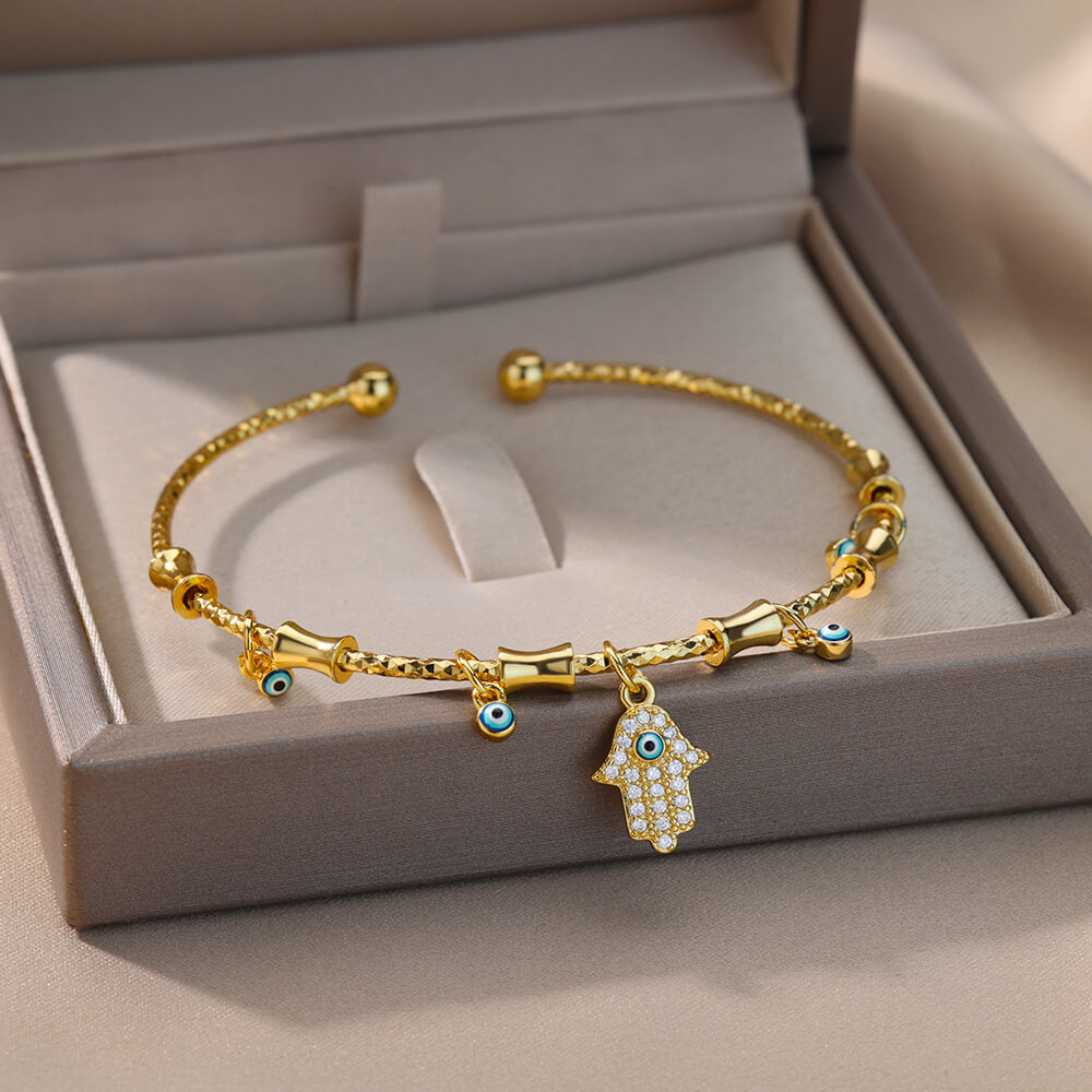 Bracelet jonc doré orné de multiples petits yeux bleus protecteurs et d'un pendentif main de Fatma serti de zircons scintillants, présenté dans un écrin.