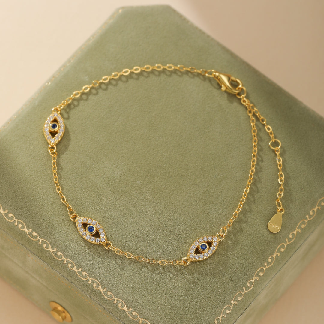 Bracelet plaqué or avec des breloques en forme d'œil maléfique ornées de zirconiums blancs et d'une pierre bleue centrale, sur une fine chaîne dorée.