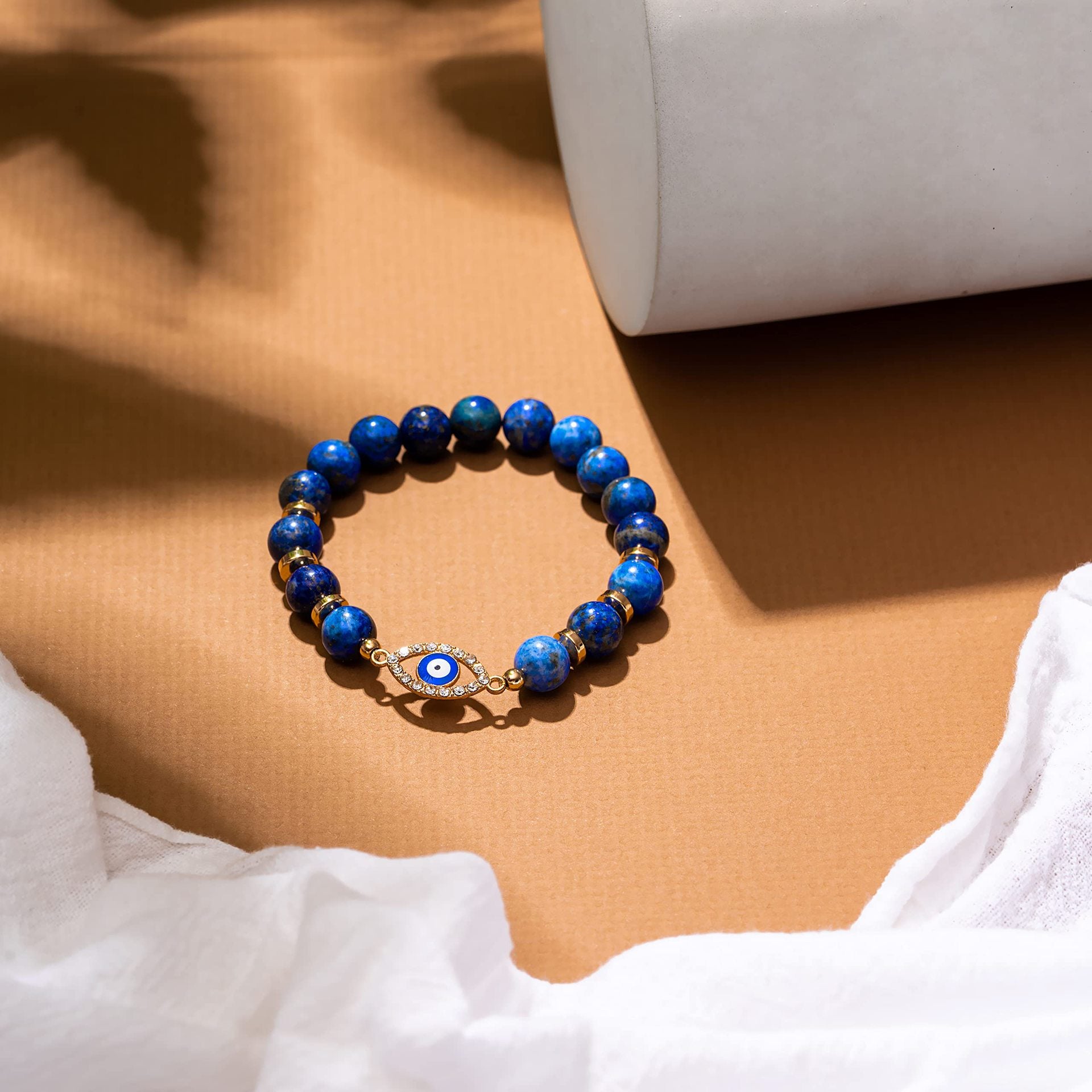 Bracelet en lapis lazuli avec un pendentif œil bleu et doré serti de strass, sur fond marron avec un tissu blanc.