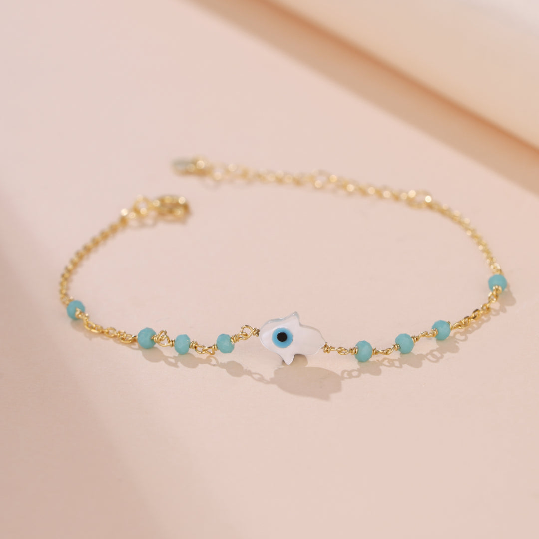 Bracelet fin en argent plaqué or avec une chaîne ornée de perles bleues turquoise et d'un pendentif main de Fatima blanc avec un œil bleu au centre, symbolisant la protection.