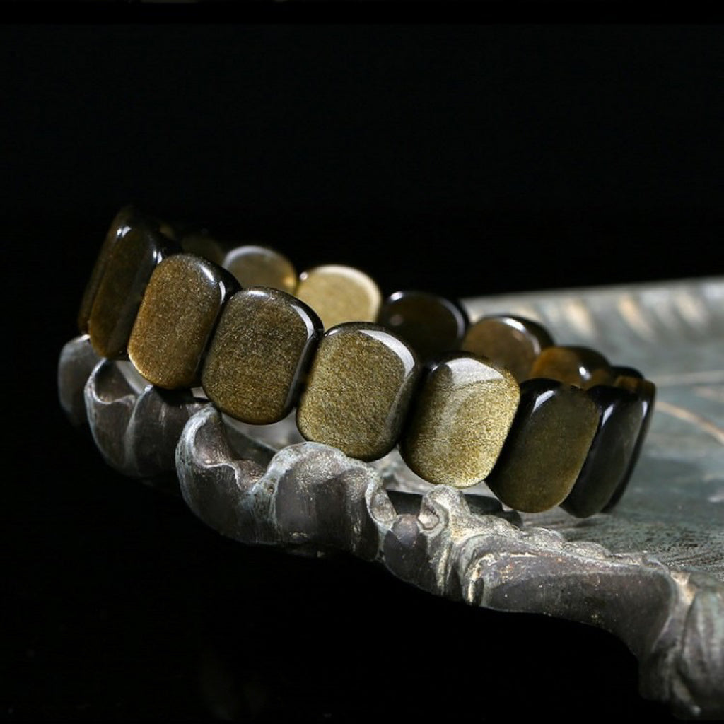 Bracelet en obsidienne dorée aux perles rectangulaires polies, posé sur un support décoratif sombre.