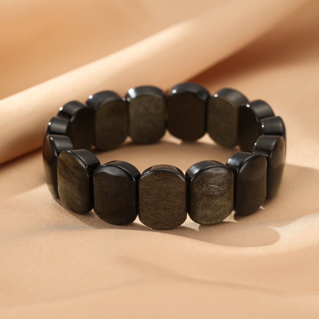 Bracelet fait de pierres d'obsidienne dorée rectangulaires, montées sur un élastique, sur un fond de tissu beige satiné.