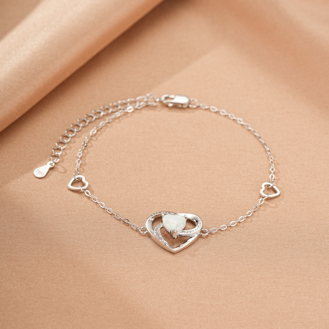 Bracelet en argent avec un pendentif en forme de cœur entrelacé orné d'une opale blanche et de petites pierres scintillantes, avec trois petits cœurs décoratifs le long de la chaîne.
