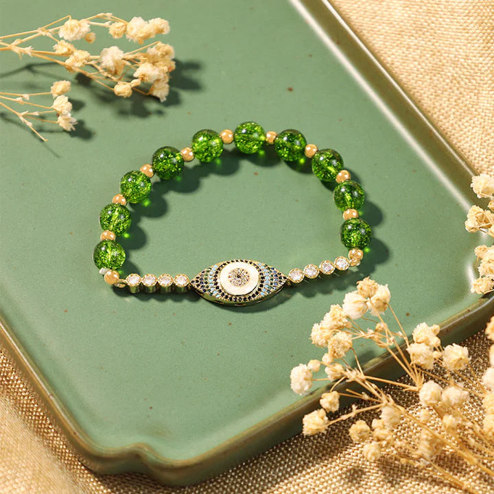 Bracelet élégant en perles de péridot vert et or, orné d'un pendentif œil bleu et blanc serti de cristaux, sur un plateau vert.