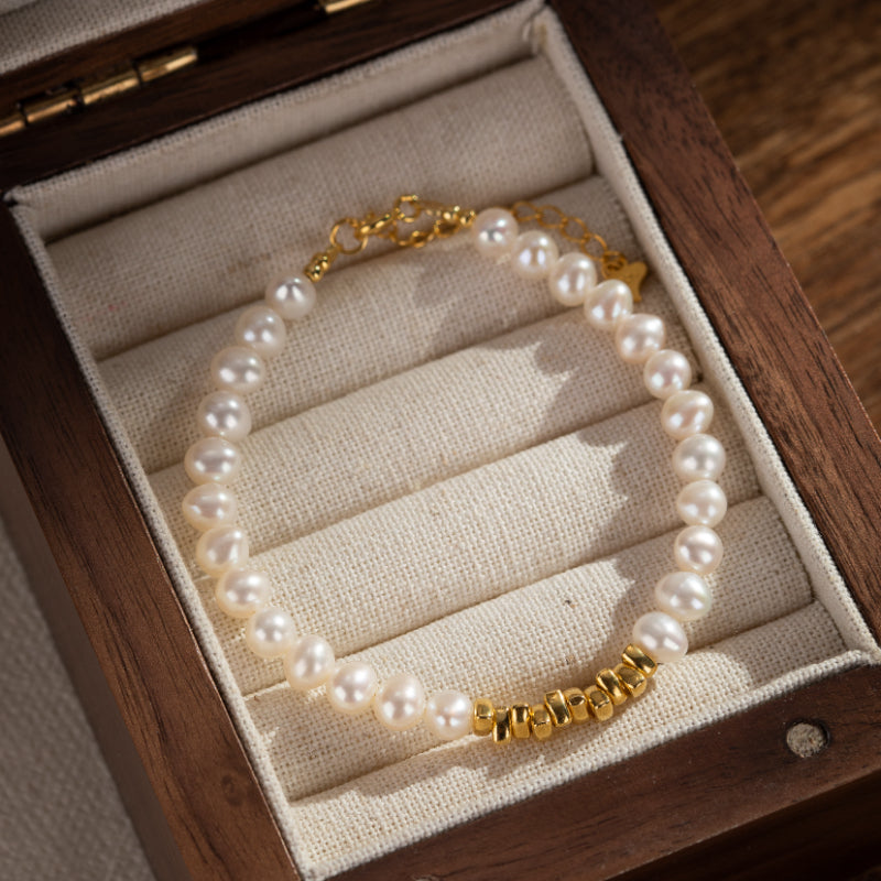 Bracelet en perles d'eau douce blanches avec des accents dorés cylindriques et un fermoir mousqueton doré, présenté dans une boîte en bois doublée de tissu.