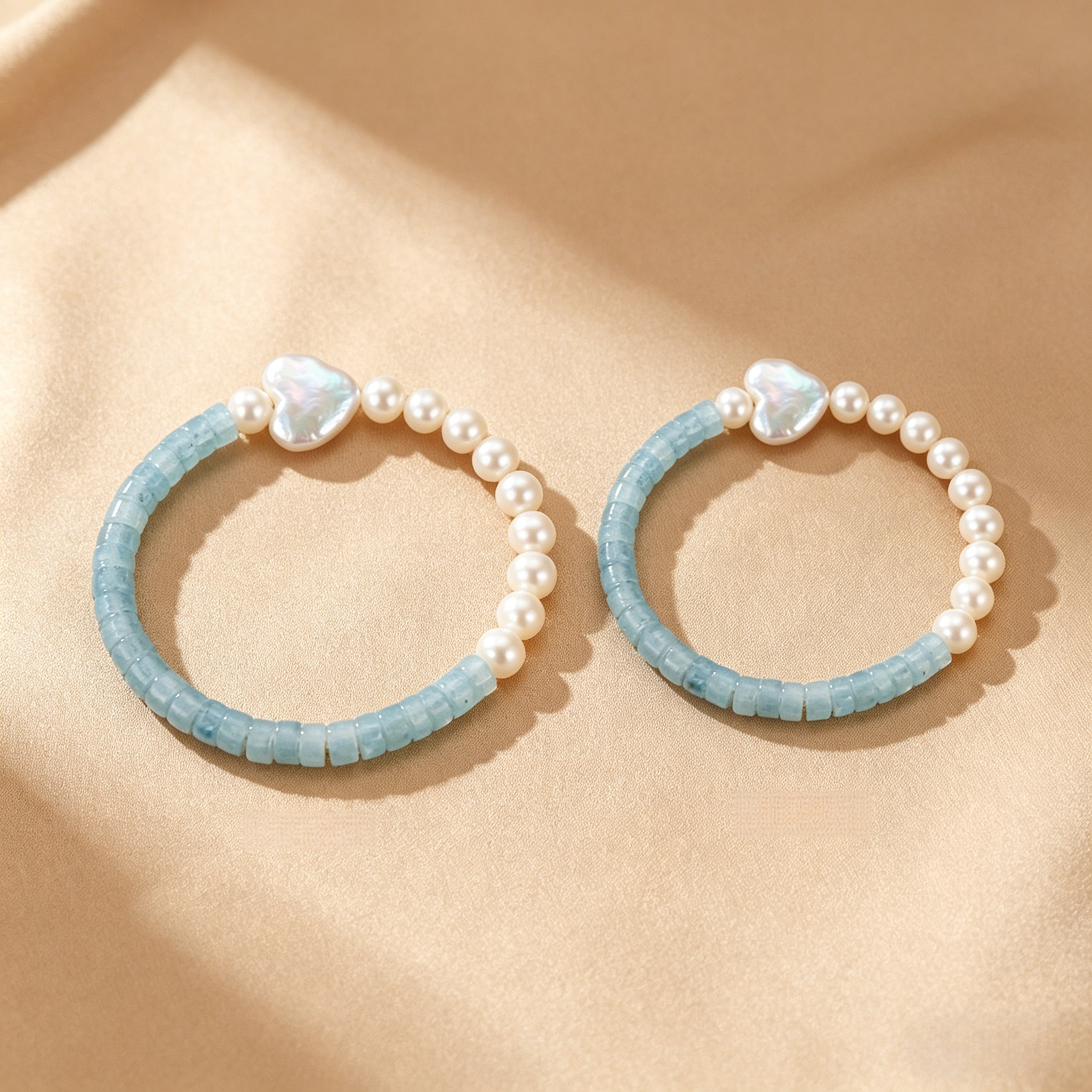 Deux bracelets en perles et aigue-marine, dont un orné d'un pendentif en forme de cœur, sur fond satiné beige.