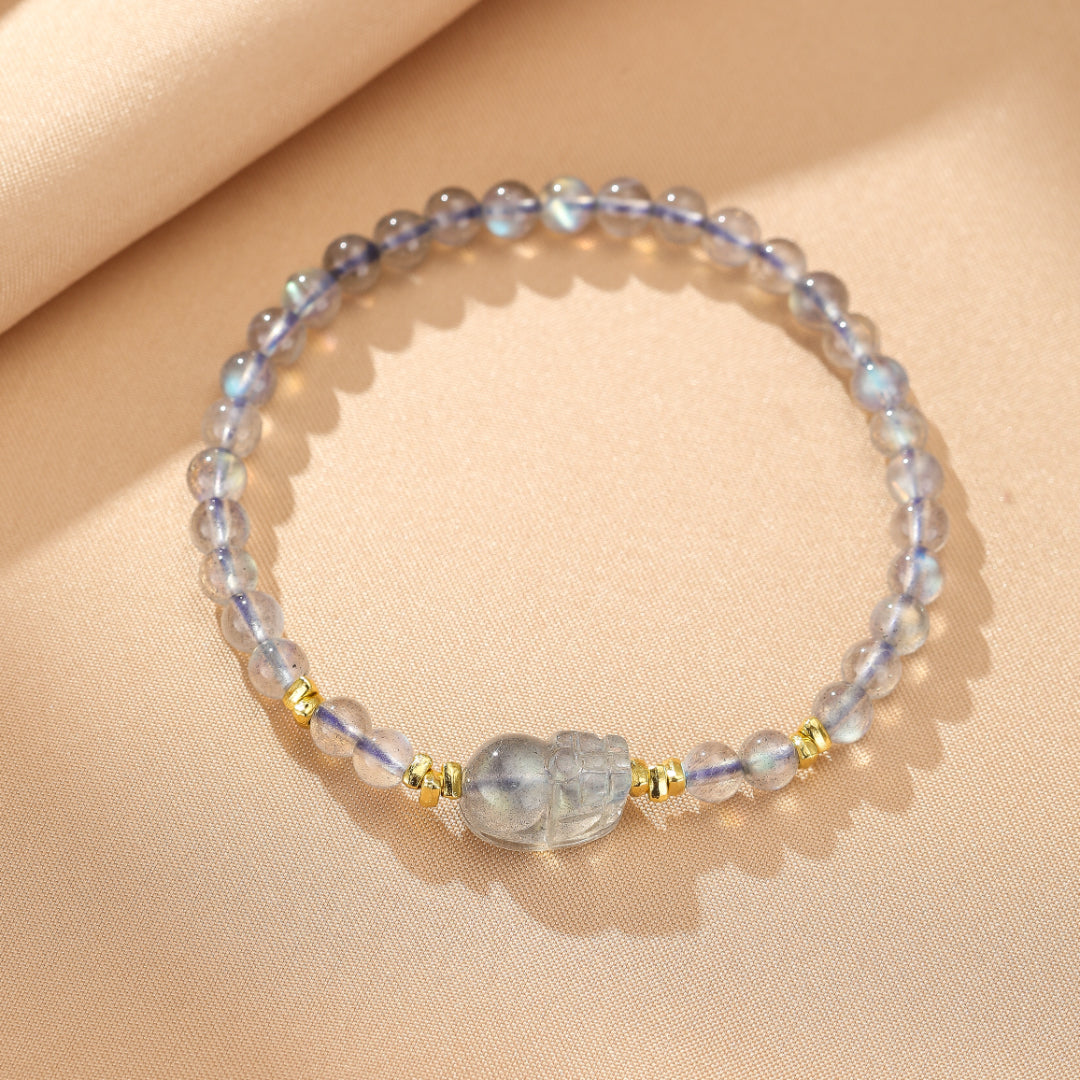 Bracelet élégant avec des perles bleues et grises de labradorite et de pierre de lune, orné d'une breloque Pi Xiu translucide et de petites perles dorées, sur un fond beige.
