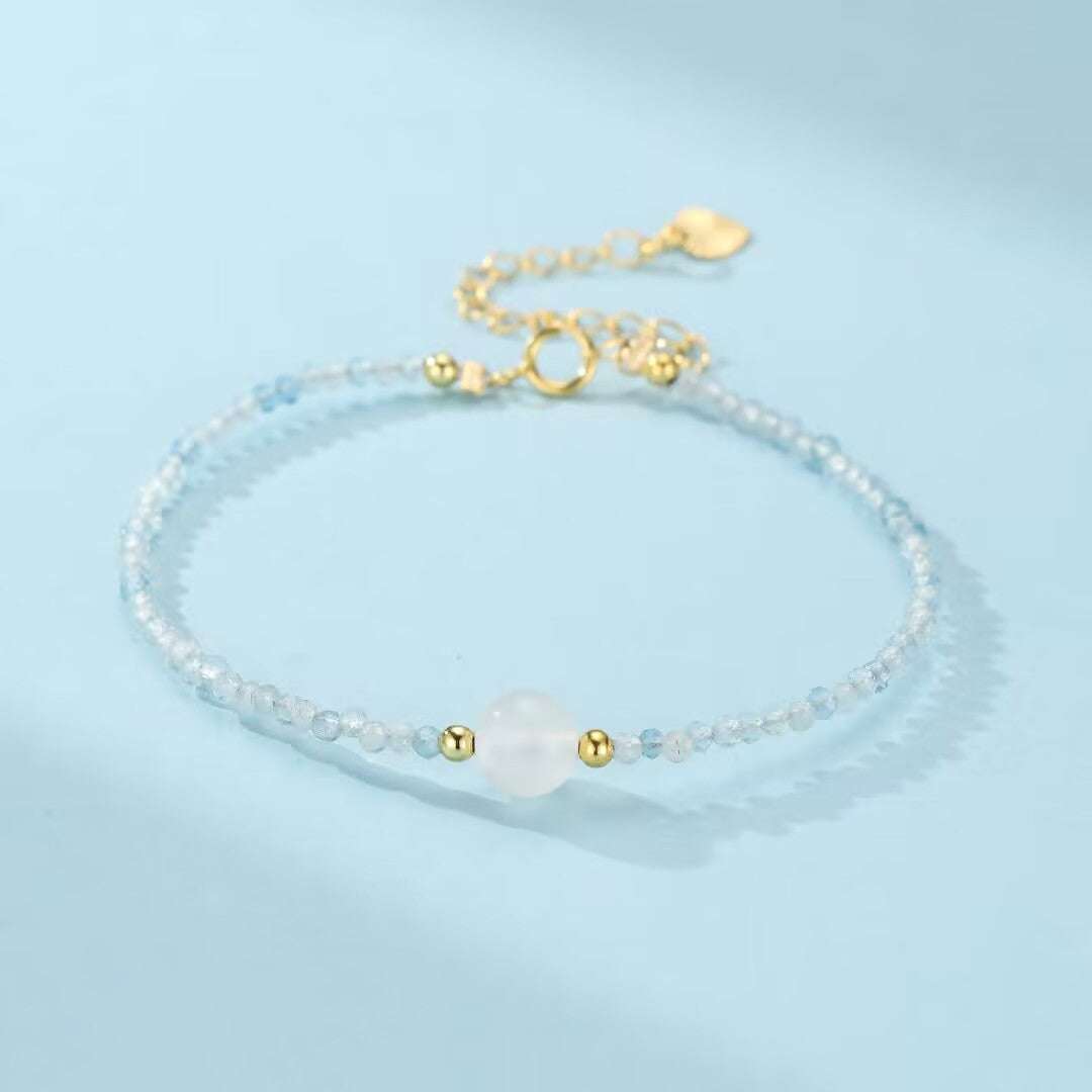 Bracelet délicat "Rêverie des flots" en pierre de lune blanche et aigue-marine bleue, avec des perles dorées, sur fond bleu ciel.