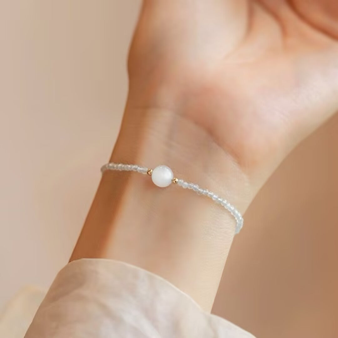 Bracelet délicat avec une pierre de lune blanche centrale et de petites perles bleues claires, porté au poignet, évoquant la douceur des vagues.
