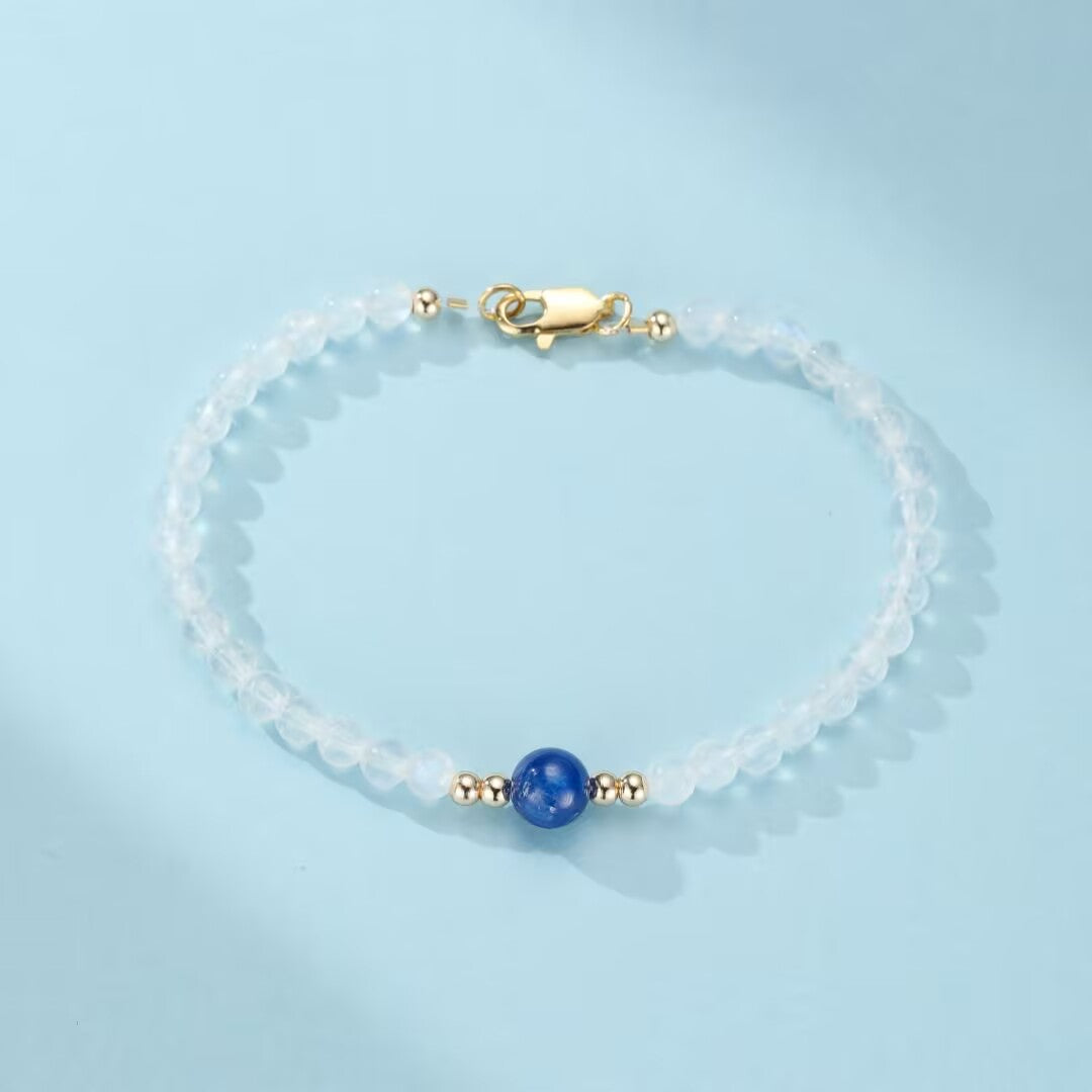 Bracelet fin en pierres de lune blanches et une perle bleue centrale, fermoir doré, sur fond bleu clair.