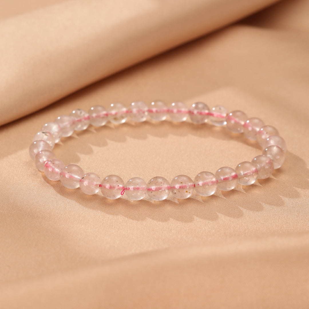 Gros plan sur un bracelet en perles de quartz fraise transparentes et roses sur un fond beige satiné. Le bracelet est disposé en courbe sur la surface.