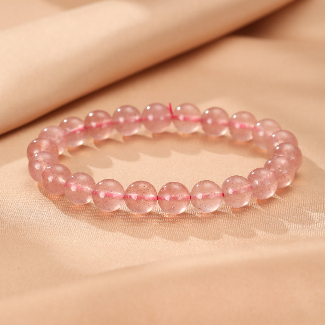 Bracelet fait de perles rondes de quartz fraise rose pâle, montées sur un fil rouge visible, posé sur un fond texturé beige clair.