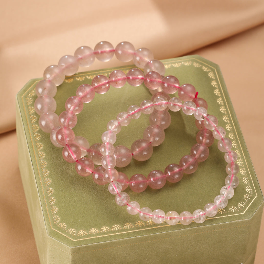 Trois bracelets en perles de quartz fraise rose clair, enfilés sur un fil rouge, reposant sur une boîte de présentation verte ornée.