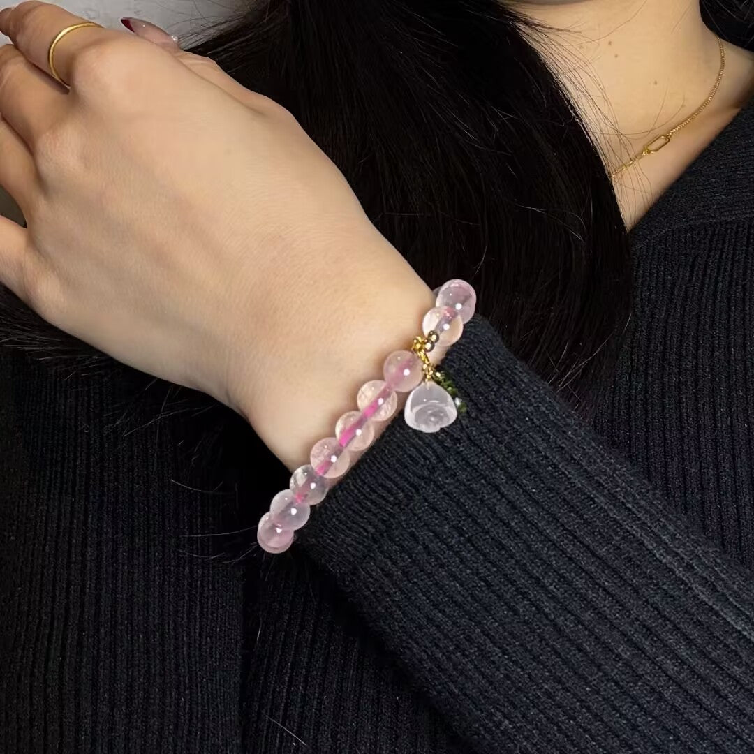 Bracelet en quartz rose avec une breloque en forme de cœur rose translucide et une petite breloque verte, porté au poignet d'une femme.