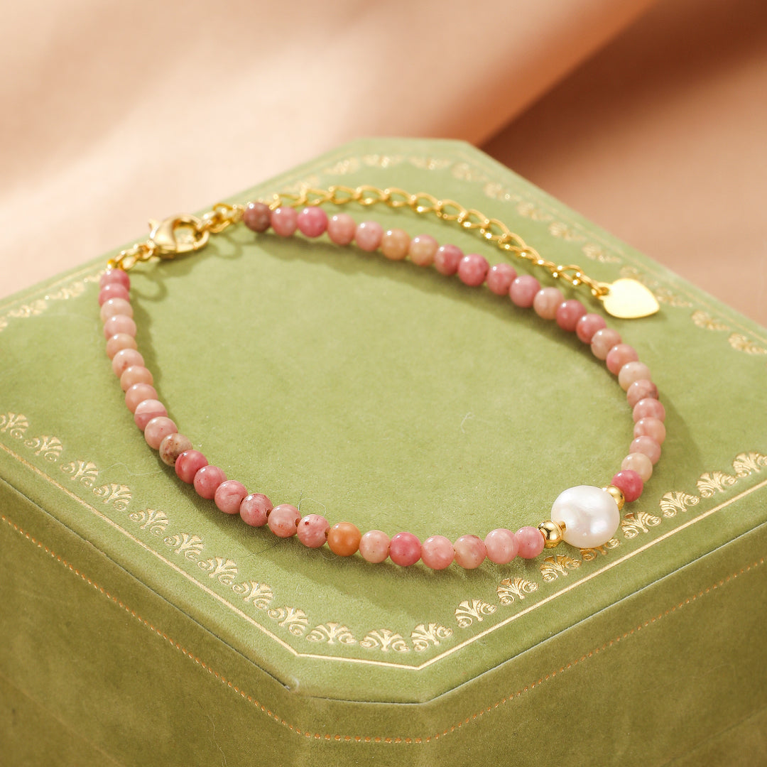 Bracelet fin en perles de rhodonite roses avec une perle blanche centrale et un fermoir doré, posé sur un écrin vert.