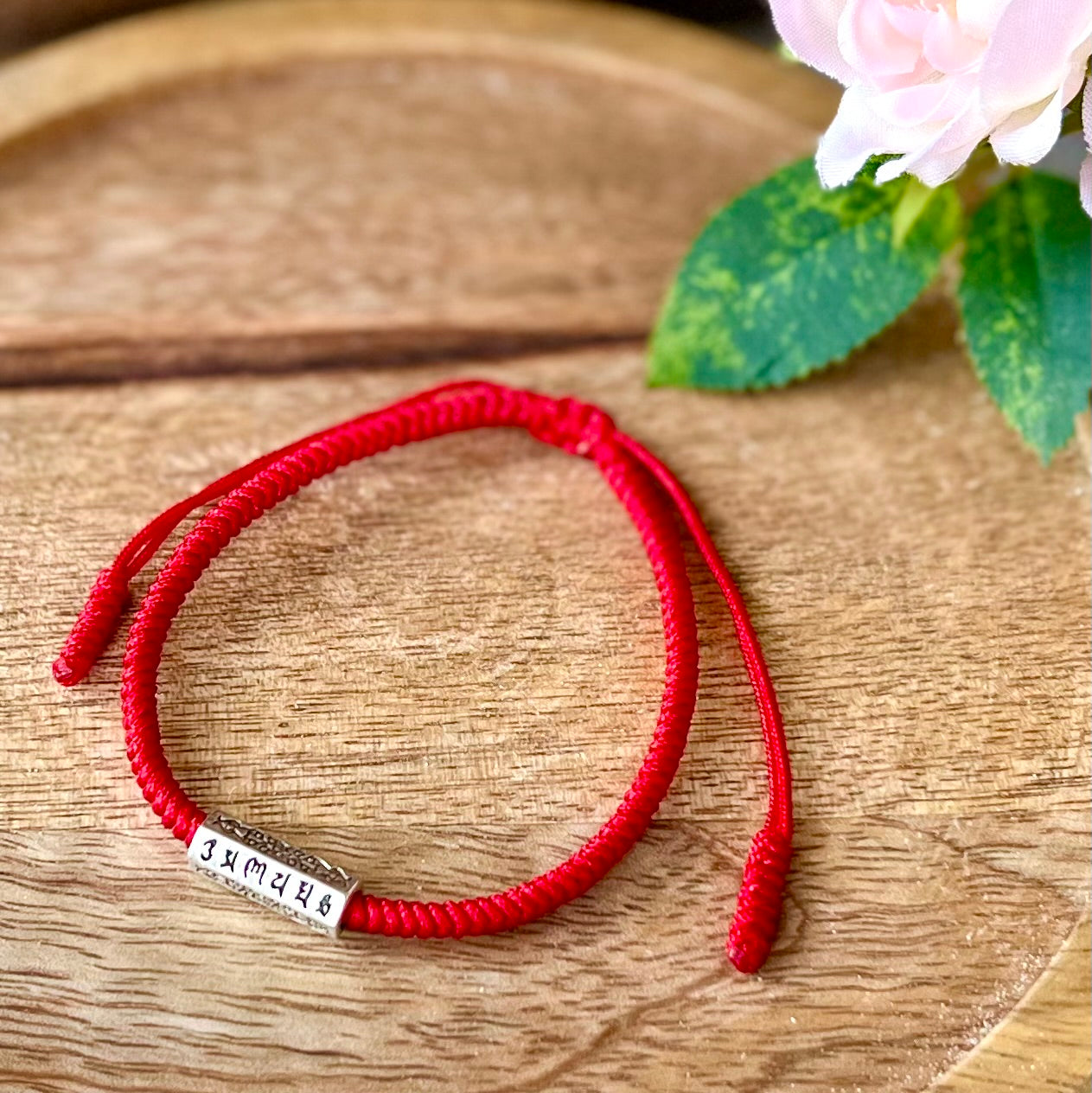 Bracelet fil rouge avec une breloque argentée gravée de six mantras bouddhistes, symbolisant l'harmonie et la créativité.