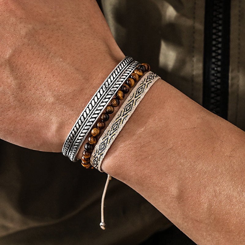 Ensemble de trois bracelets pour homme : un bracelet en argent gravé, un bracelet de perles œil de tigre et un bracelet en tissu à motif ethnique.