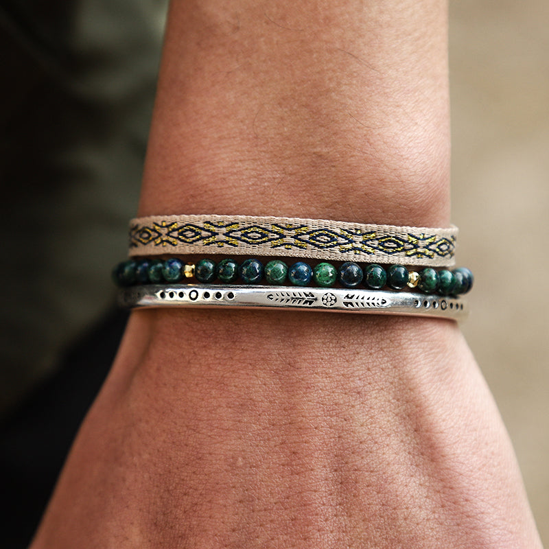 Ensemble de trois bracelets ethniques portés au poignet : un bracelet tissé beige et noir, un bracelet de perles vertes et un bracelet rigide argenté gravé.