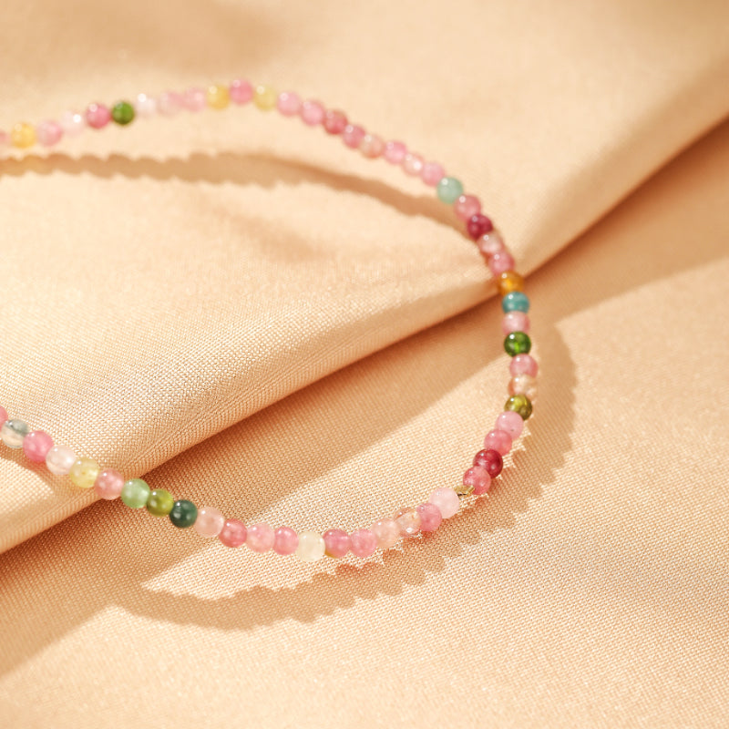 Bracelet fin en tourmaline multicolore, perles fines roses, vertes et bleues, sur un fond de tissu satiné beige.