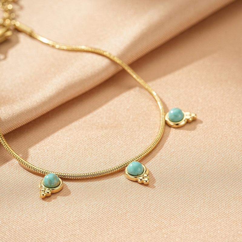 Bracelet chaîne dorée ornée de trois petites pierres turquoise rondes serties sur des montures dorées en forme de goutte, posé sur un tissu beige.
