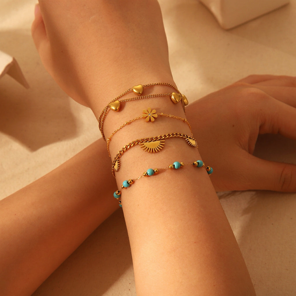 Bracelet chaîne doré avec perles turquoise et breloques soleil, cœur et fleur, porté sur un poignet.