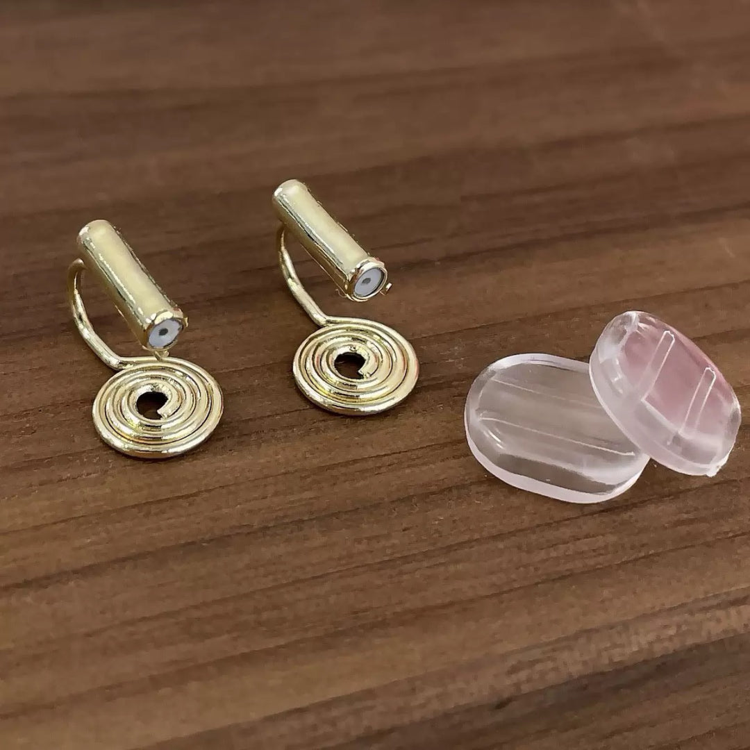 Paire de clips d'oreilles dorés en forme de spirale élégante, avec un petit cylindre sur le dessus et deux coussinets d'oreille transparents à côté, le tout sur un fond en bois.
