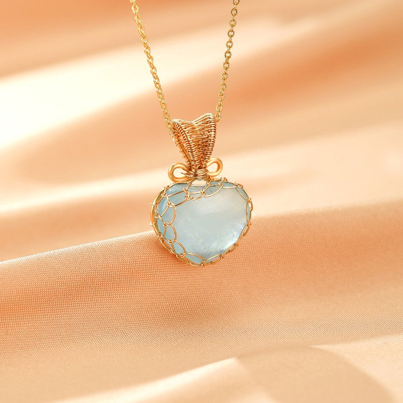 Collier en or jaune avec un pendentif cœur en aigue-marine entouré de fils d'or tressés, sur fond satiné couleur pêche.