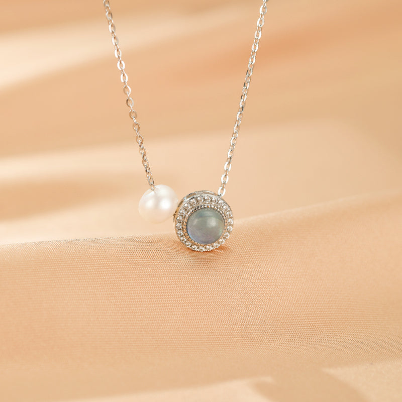 Collier en argent 925 avec une aigue-marine ronde sertie de petits cristaux et une perle blanche nacrée, sur fond satiné pêche.