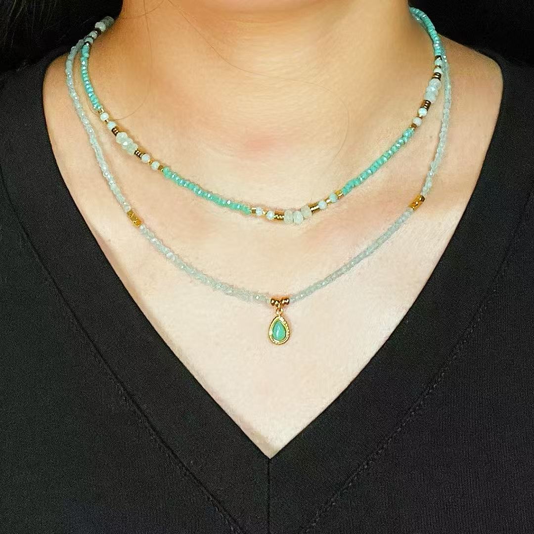 Trois colliers superposés : deux colliers fins sertis de petites perles turquoise et dorées, et un collier plus fin avec un pendentif en forme de goutte vert émeraude.