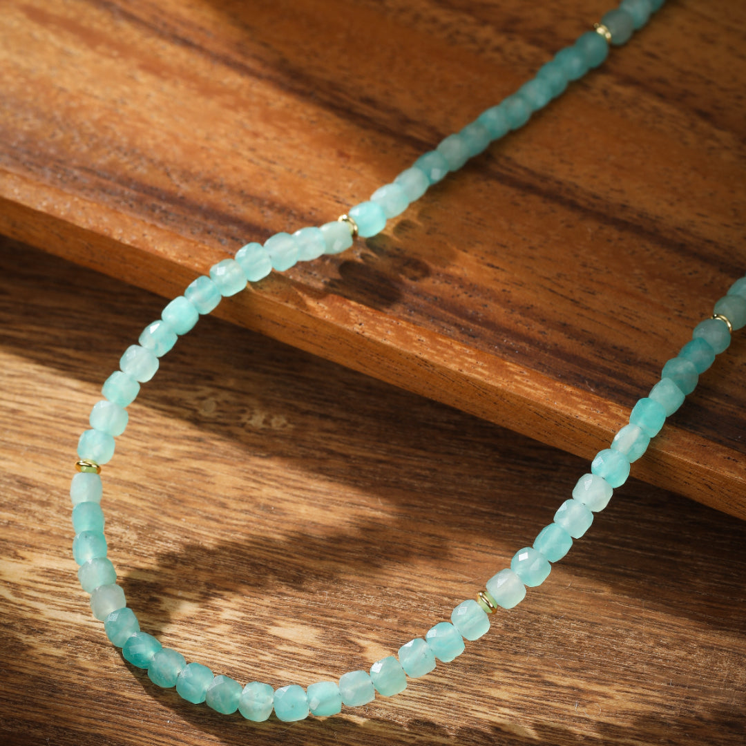Collier fin en amazonite aux perles facettées bleu pâle, rehaussé de petites perles dorées, posé sur un fond en bois sombre.