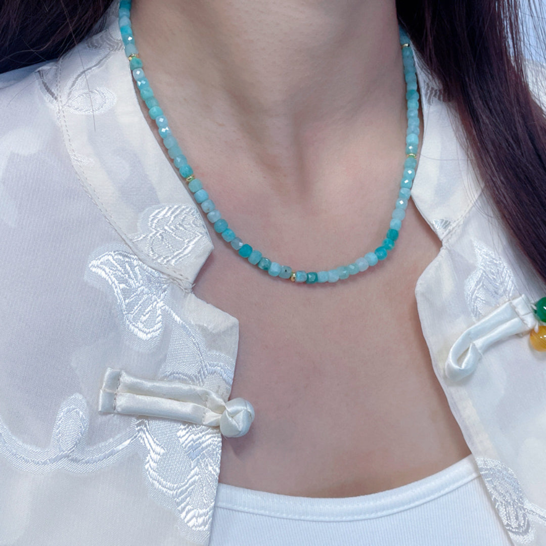 Gros plan sur un collier délicat en amazonite aux teintes bleu clair et turquoise, ponctué de petites perles dorées, porté par une femme portant une chemise blanche.