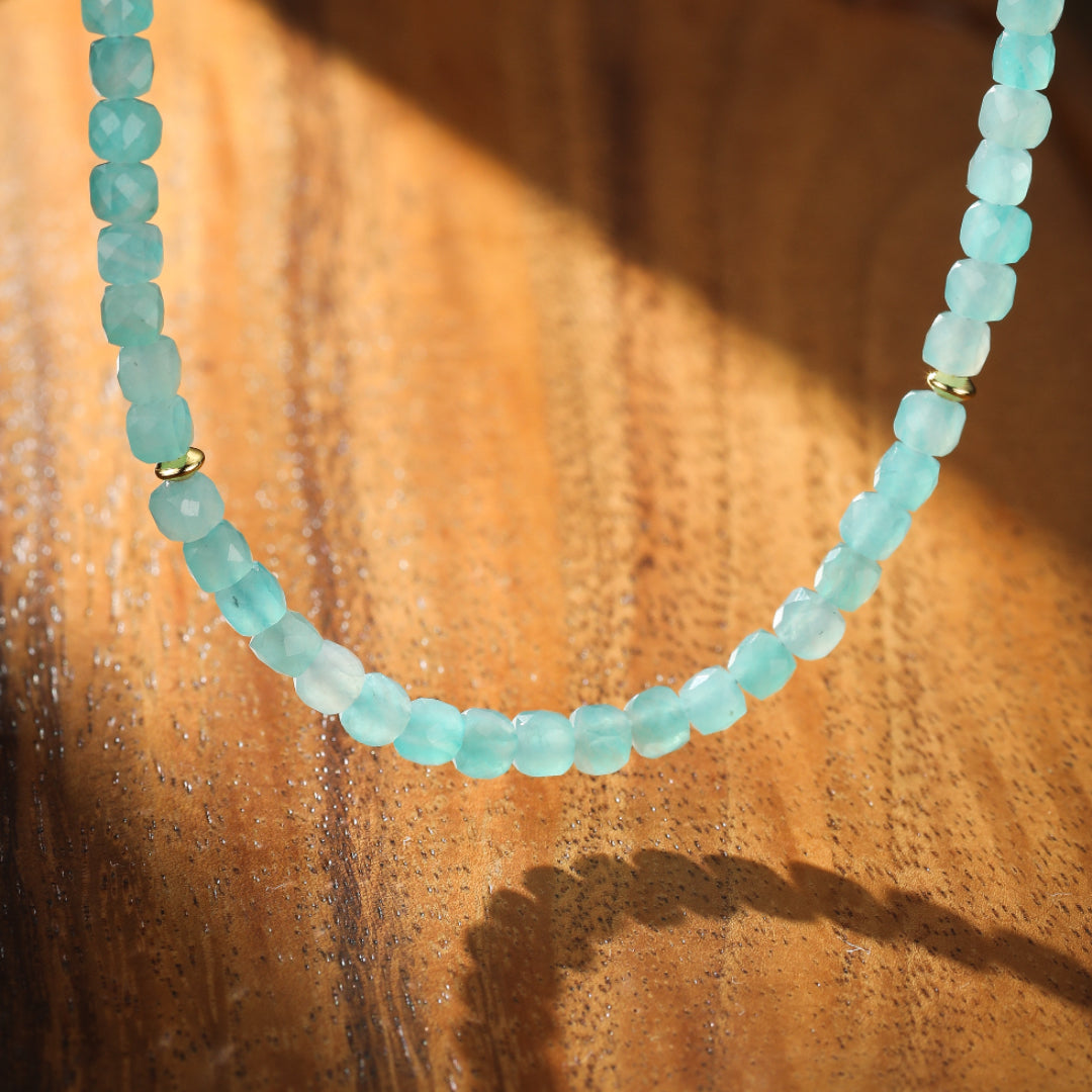 Collier fait de petites pierres d'amazonite facettées bleu-vert pâle, avec une petite perle dorée au centre, posé sur un fond en bois avec des ombres.