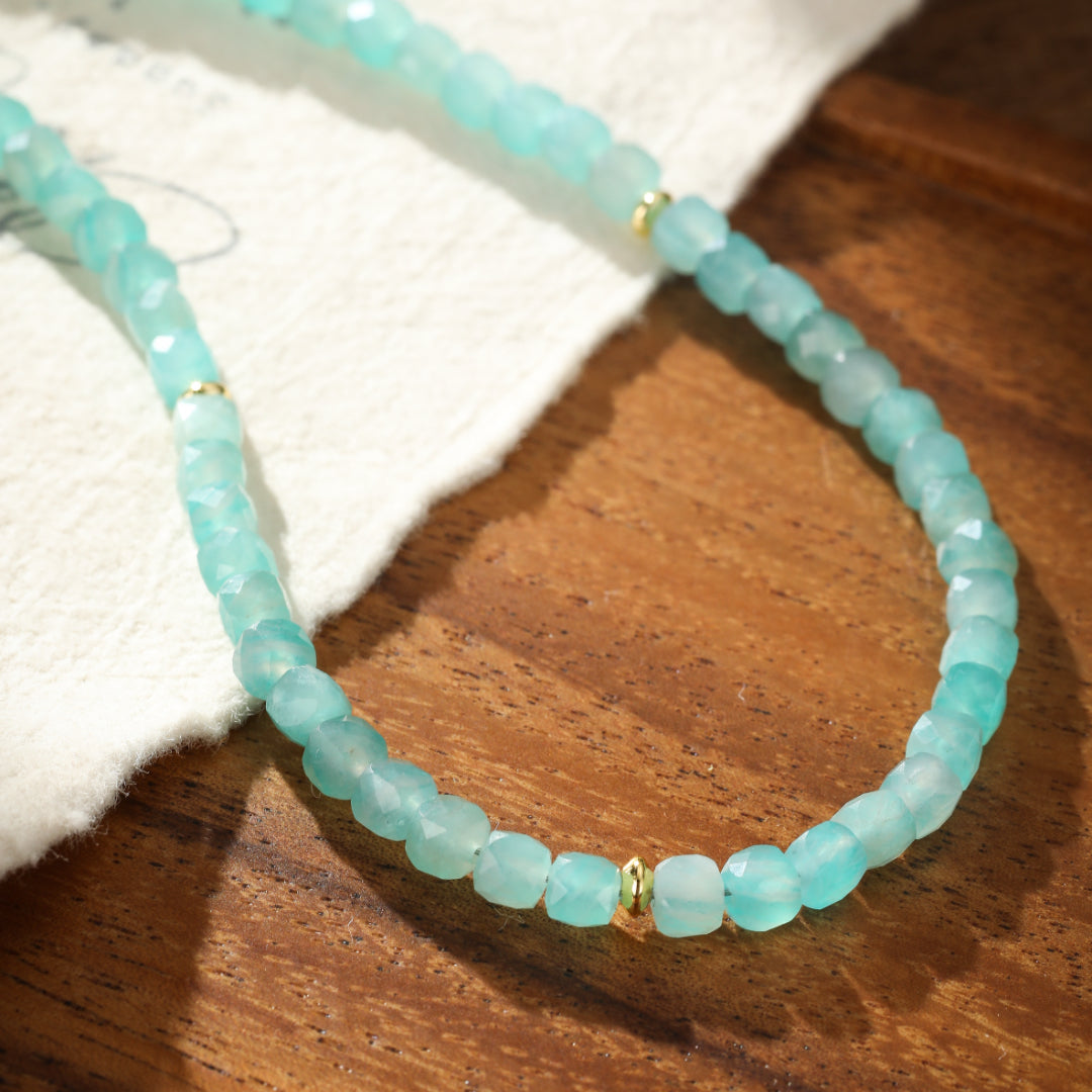 Collier fait de petites pierres d'amazonite bleu-vert facettées et de perles dorées, posé sur un tissu blanc et un fond en bois.