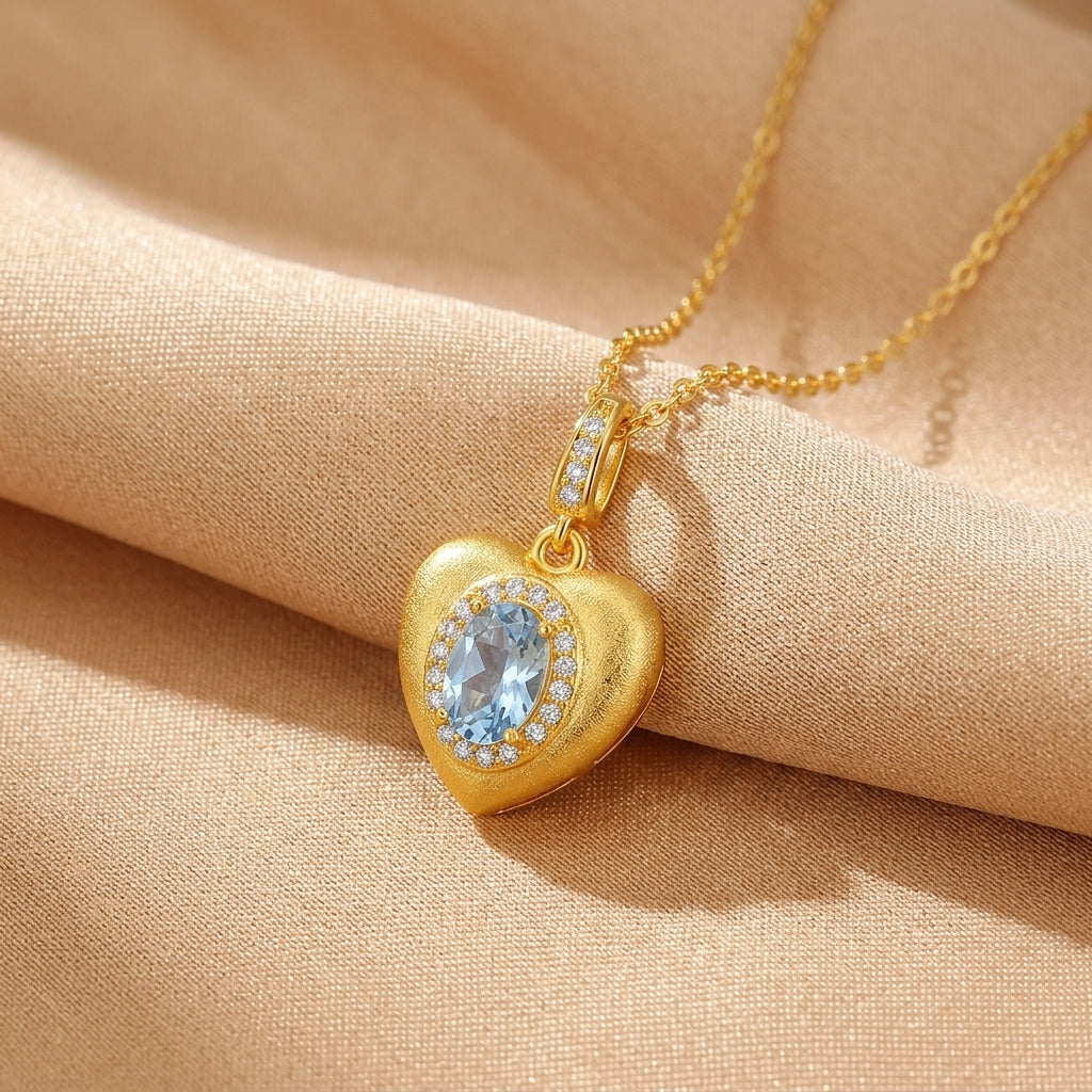 Collier en forme de cœur en argent plaqué or avec une pierre bleue ovale au centre, entourée de petites pierres scintillantes, sur un fond de tissu beige.
