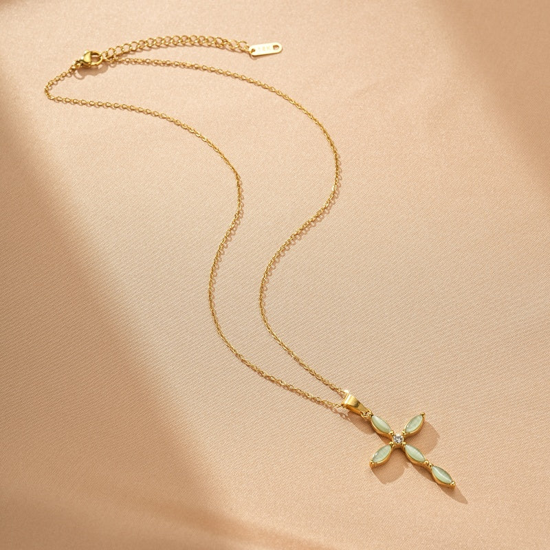 Collier doré avec un pendentif en forme de croix orné de pierres vertes et d'un petit diamant au centre, sur fond beige.