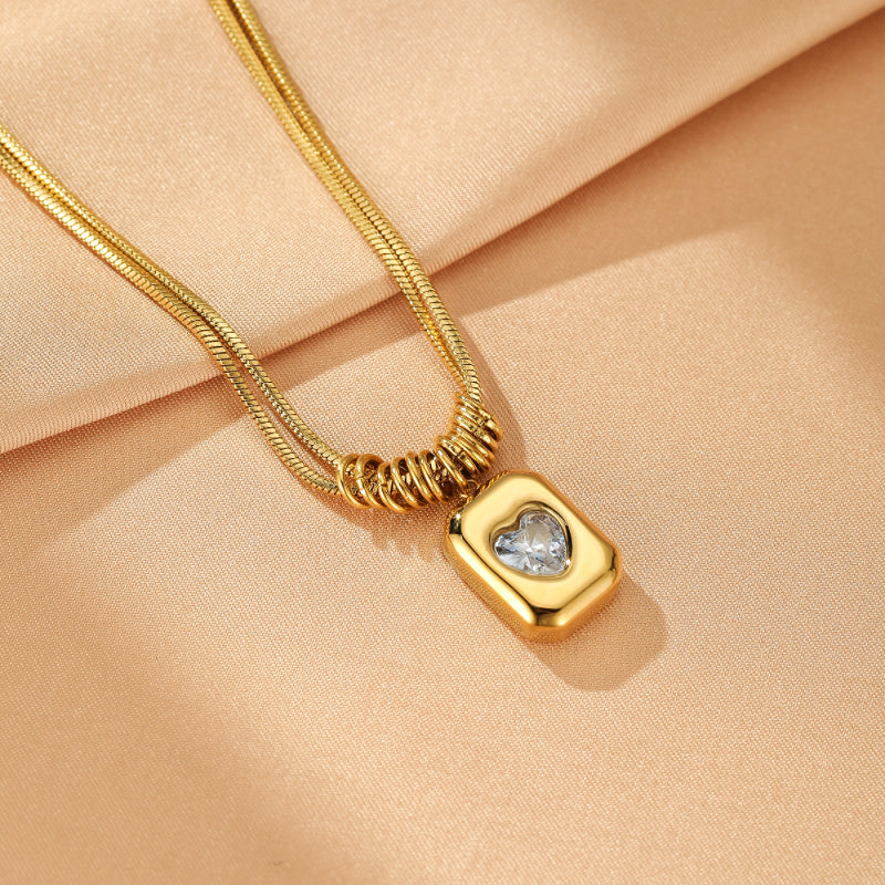 Collier Cœur Rayonnant en plaqué or, pendentif rectangulaire doré avec un cœur en cristal scintillant incrusté, sur fond satiné beige.